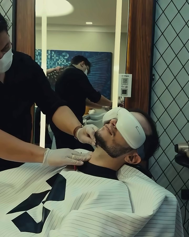 Cliente relaxando com máscara nos olhos enquanto o barbeiro cuida da barba